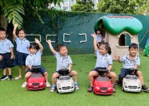 Happy children in school uniforms sitting on toy cars in a playground, with excited expressions as they raise their hands - Starshine Montessori