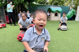 Joyful preschool children riding toy cars during outdoor playtime at Starshine Montessori preschool, supervised by a teacher in a green garden setting.