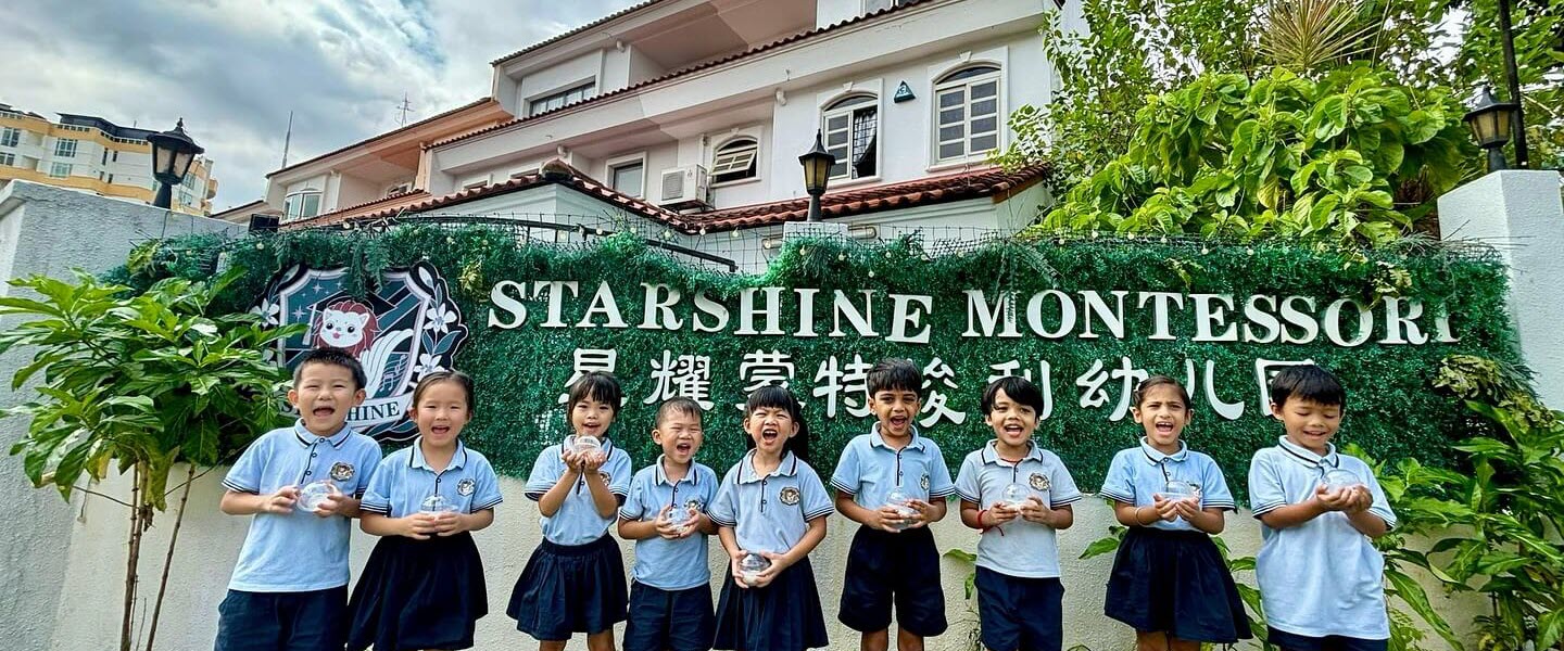 Group of happy students standing outside Starshine Montessori Preschool, smiling and holding crafts in front of the school sign. The image showcases a nurturing early childhood education environment focused on joy, community, and Montessori values.