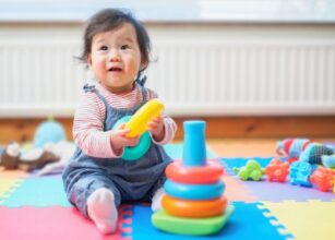 Smiling baby playing with colorful stacking rings on a foam play mat in a bright indoor environment, encouraging early learning and motor skill development.