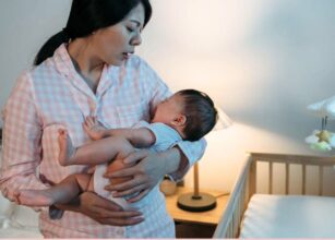 Mother in pink pajamas gently holding and soothing her baby near a crib at night.