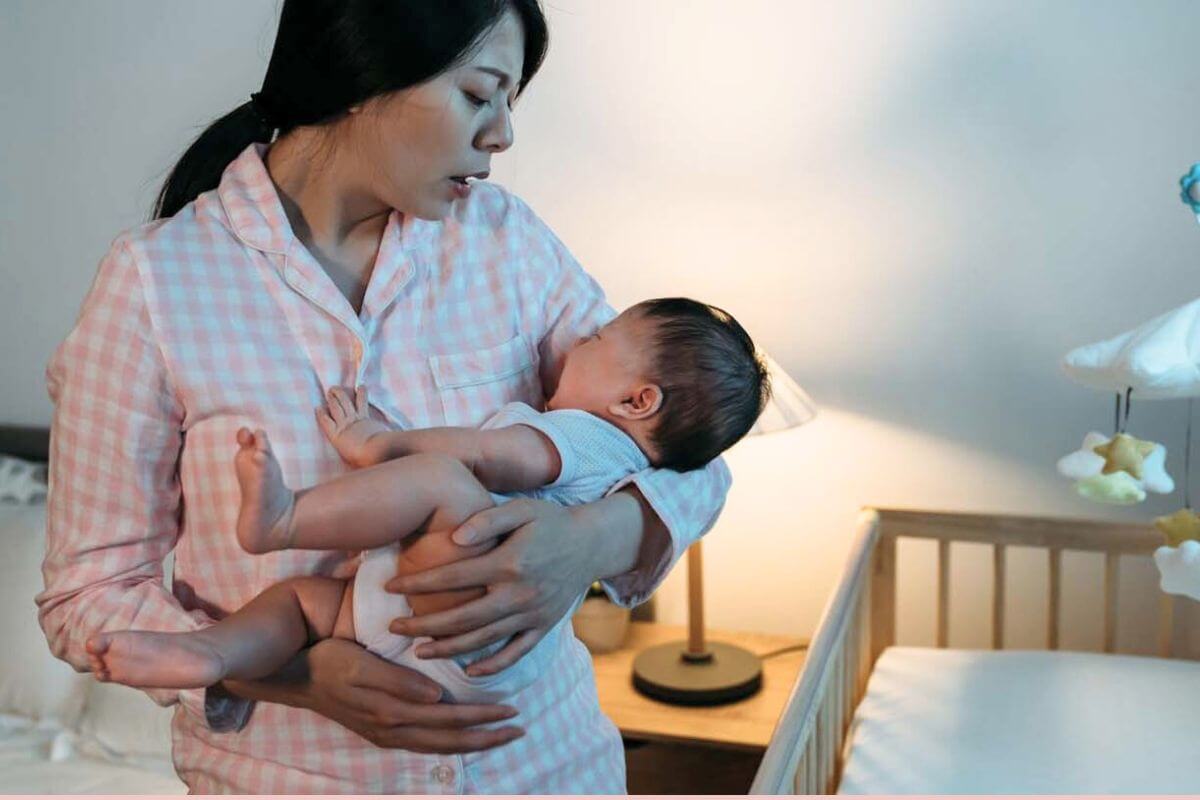 Mother in pink pajamas gently holding and soothing her baby near a crib at night.