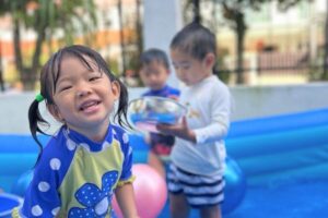 Smiling preschool girl in blue polka dot rash guard enjoying water play in an inflatable pool with other children at Starshine Montessori Hillview.