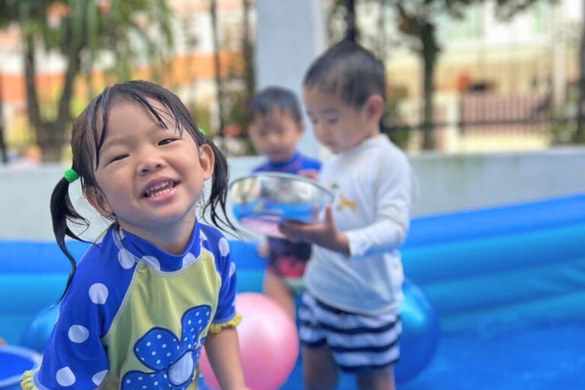 Smiling preschool girl in blue polka dot rash guard enjoying water play in an inflatable pool with other children at Starshine Montessori Hillview.